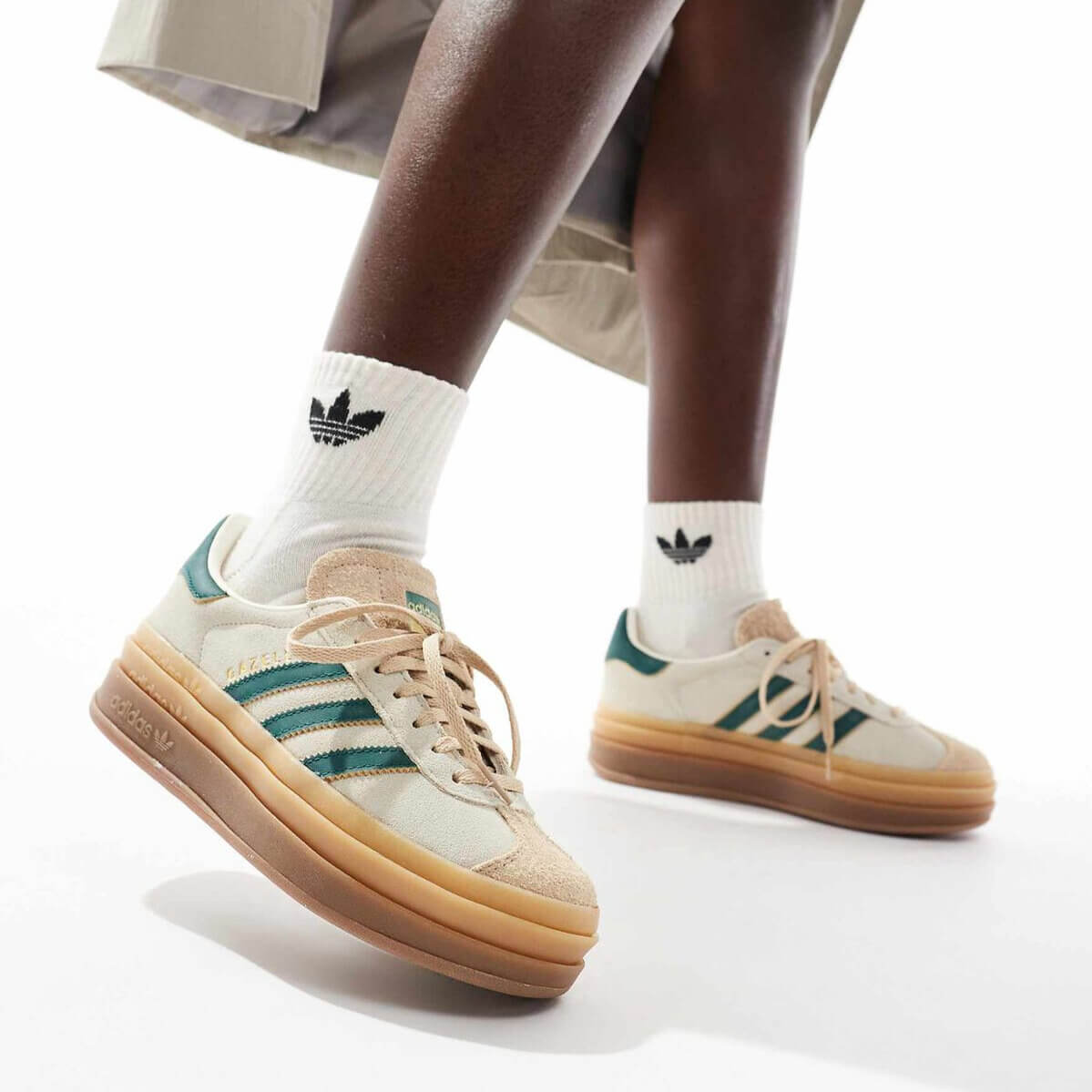 A woman wearing a khakhi skirt and brown and green Adidas Gazelle Bold sneakers